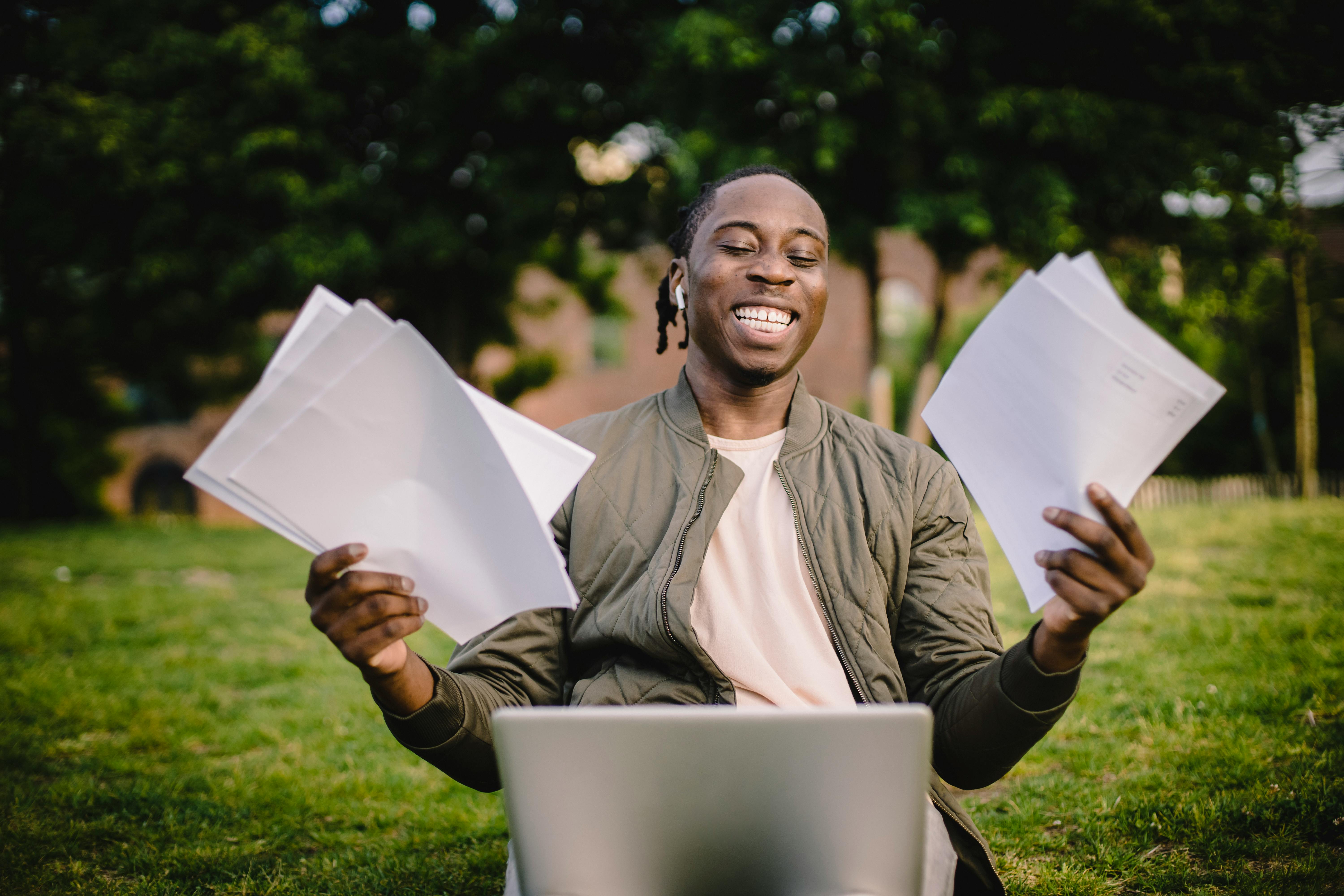 A young man sits smiling at a laptop outside whilst holding pieces of white paper. In the background grass and trees can be seen.