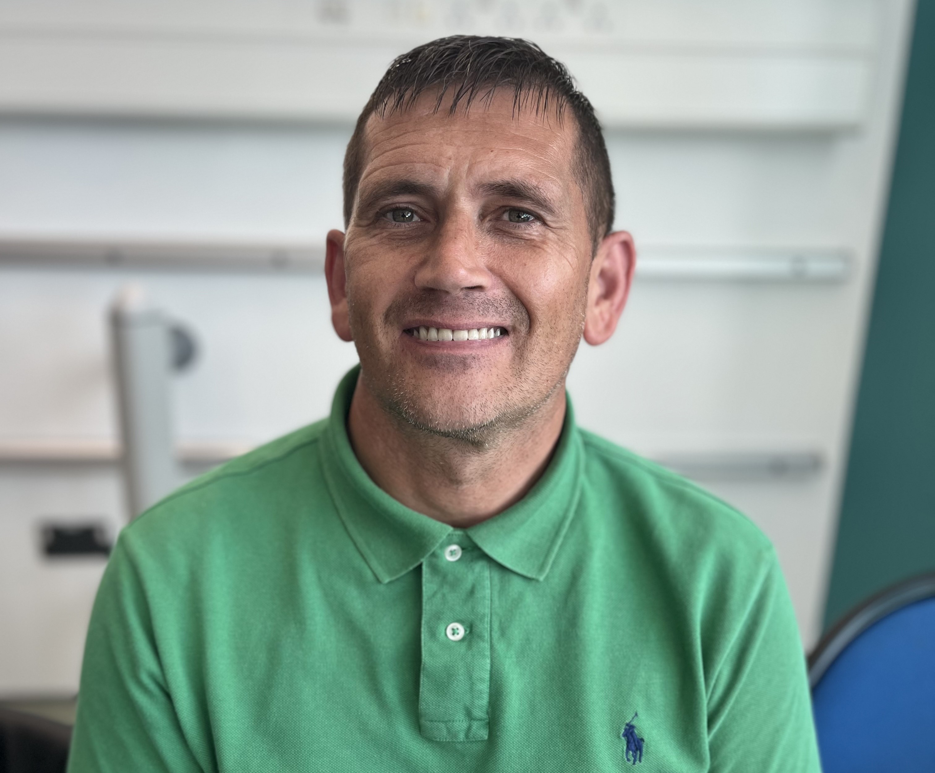 A man smiling against a blurred background. He is wearing a green polo shirt.