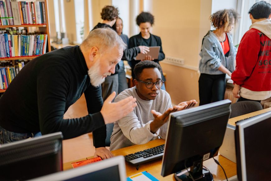 An instructor leans over a desk to help a student seated at a desktop computer in a computer lab or library. Bookshelves filled with books line the left side of the room, while several other people work and talk in the background. Multiple computer monitors are visible in the foreground, and natural light streams in through large windows, creating a bright learning environment.